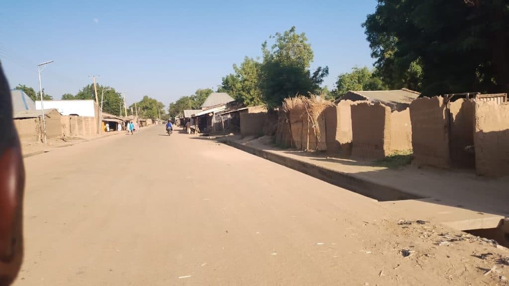 Street scene with dirt road, a few people, and mud-brick buildings under a clear blue sky.