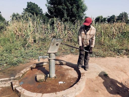 Person using a hand pump at a well in a rural area, surrounded by plants and dry ground.