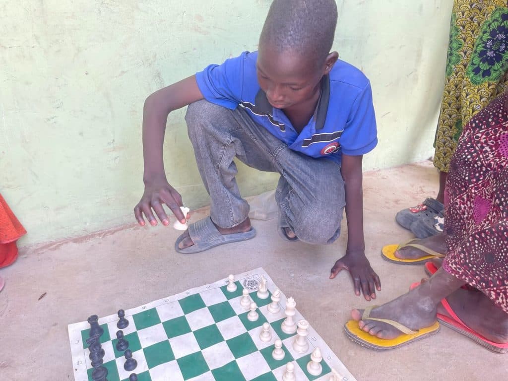 Child in a blue shirt playing chess on the floor with green and white board, surrounded by people in colorful clothing.