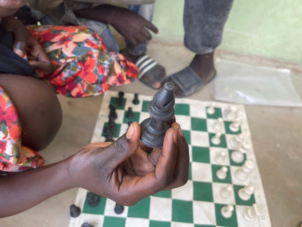 A person holds a black bishop piece over a chessboard. Two people are seated nearby, one wearing floral pants and the other in jeans and slippers.