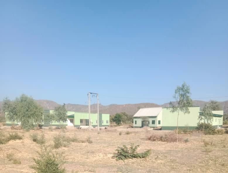 Green buildings under a clear blue sky, with dry grass and scattered trees in the foreground. Hills are visible in the background.