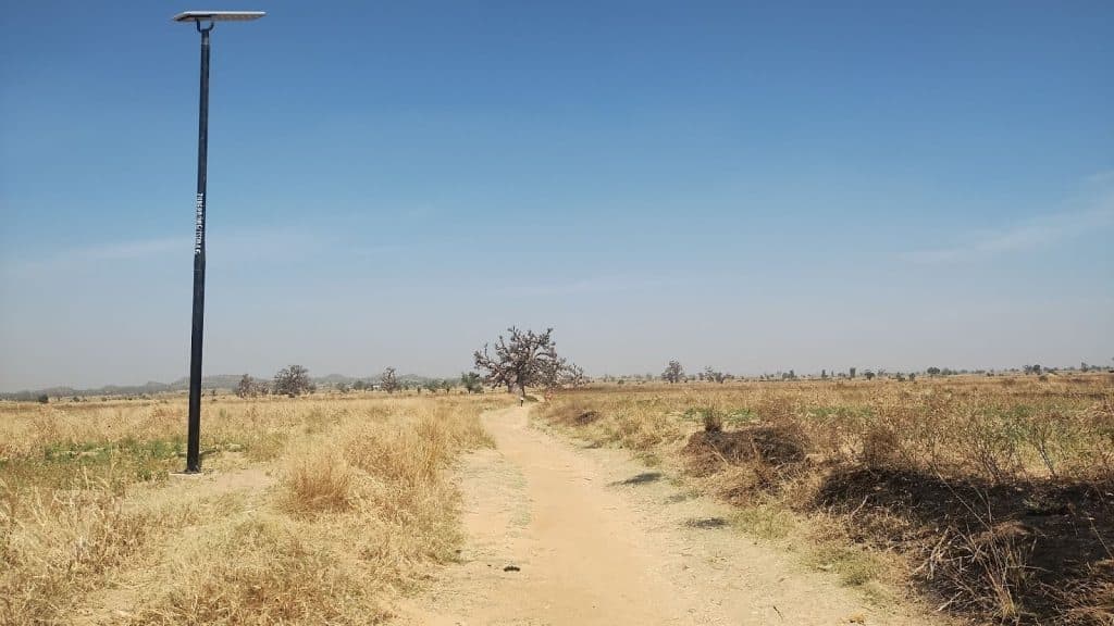 Dirt path through dry grassy field, with a solar streetlight and a distant tree under a clear blue sky.