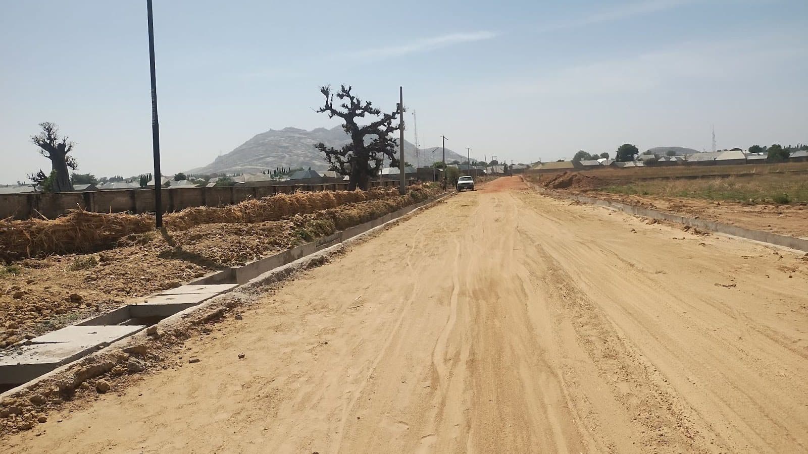 A dusty rural road with sparse trees, a parked car, and distant hills under a clear blue sky.