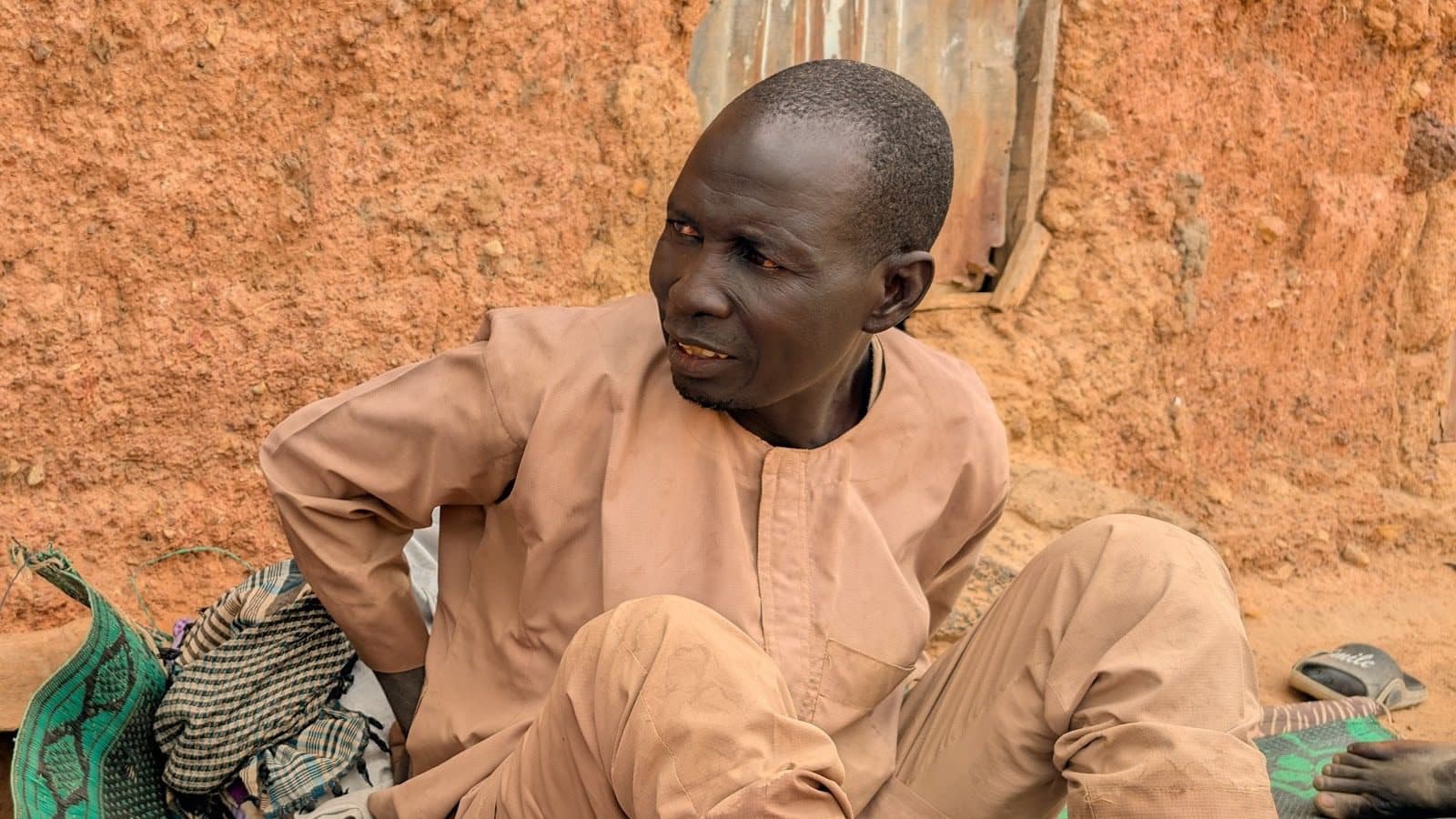 A person sitting outdoors against a textured wall, dressed in brown traditional attire, looking to the side.