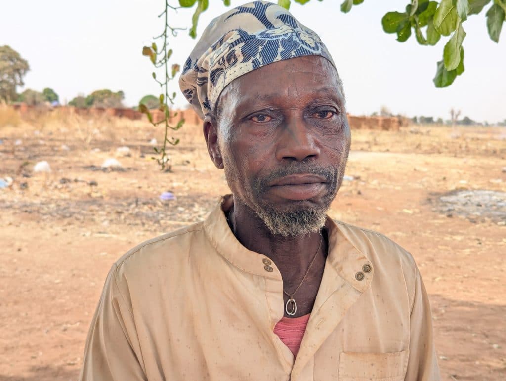A man with a patterned headscarf stands in a dry, open landscape under a leafy branch.