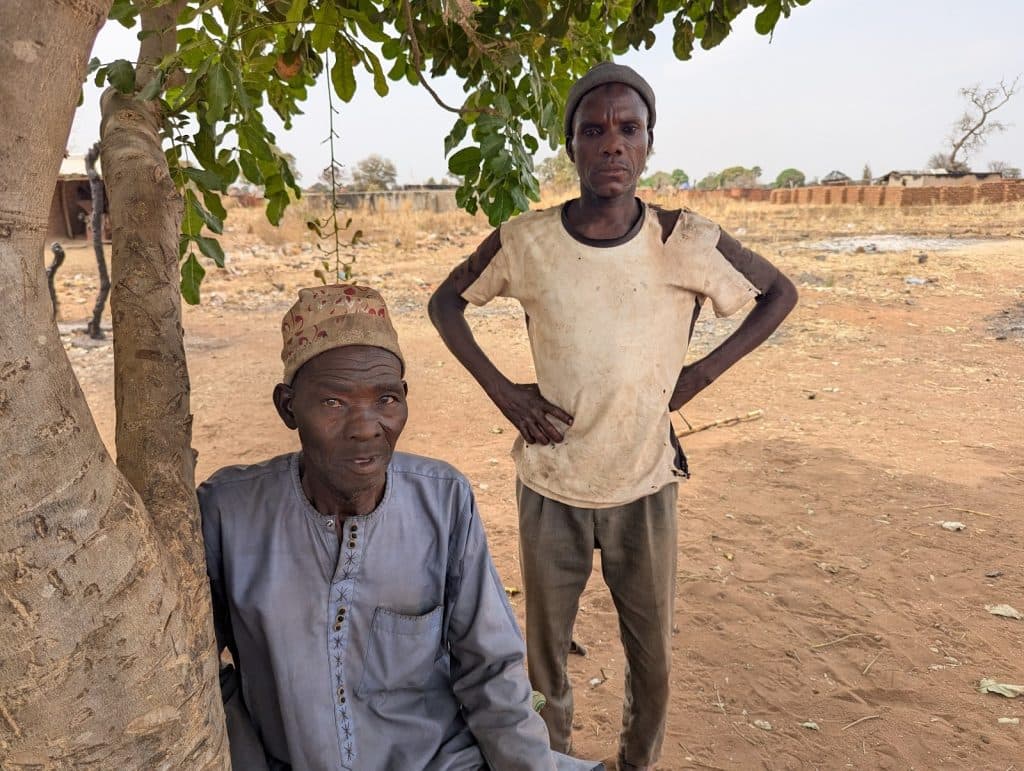 Two men outdoors in a rural area, one sitting under a tree and the other standing with hands on hips. Dry landscape in the background.