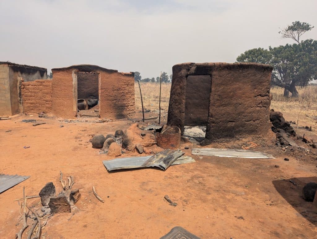 Charred remains of mud huts in a dry field, with debris and a few trees in the background.