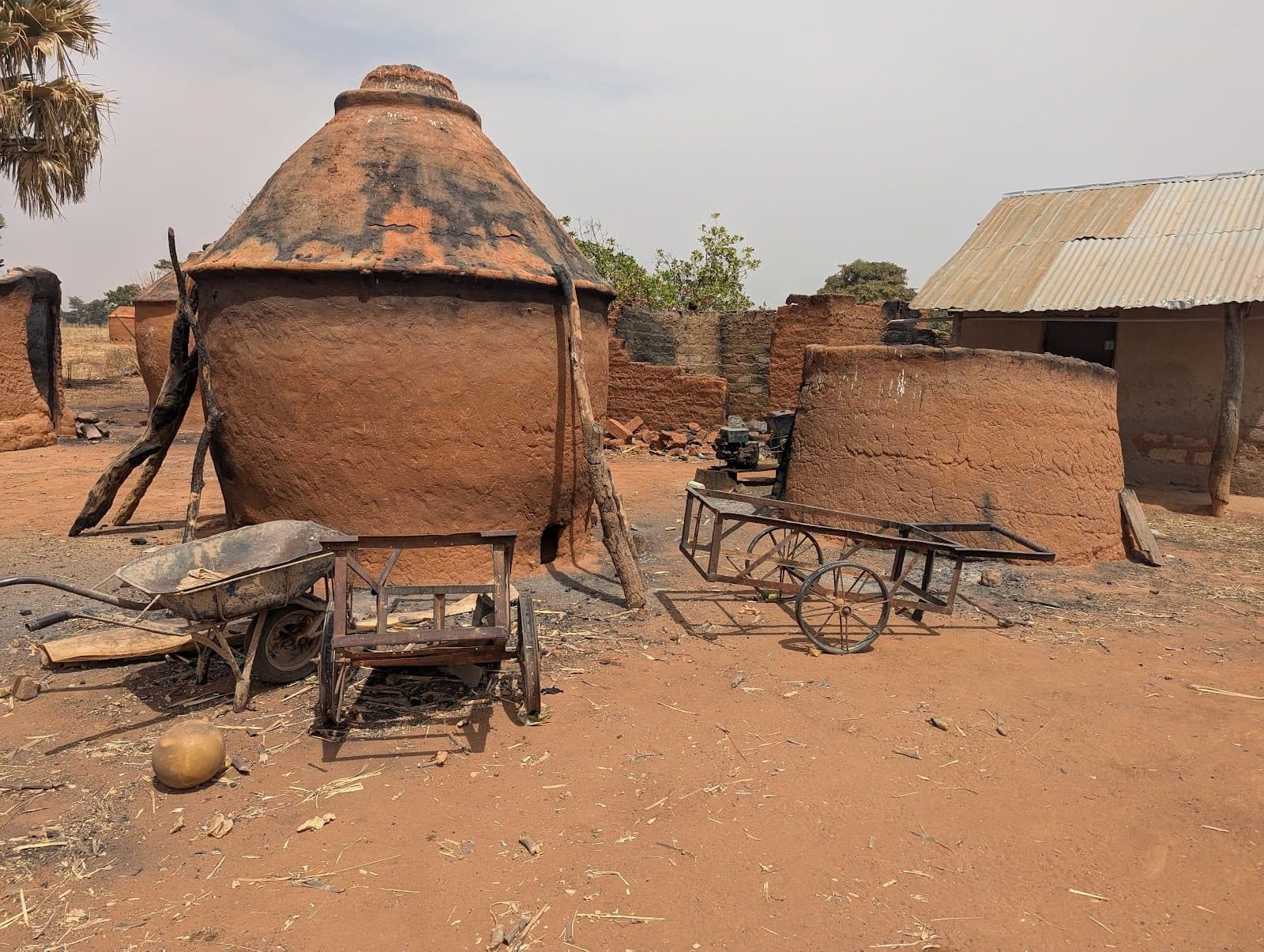 Large clay ovens with wooden supports, plus a wheelbarrow and cart in a sunlit rural setting.