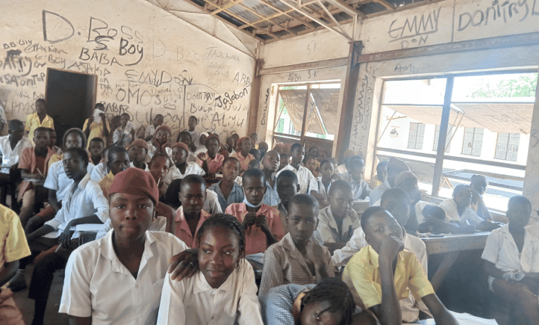 A crowded classroom with students in uniforms sitting closely, surrounded by walls covered in graffiti under a partially open roof.