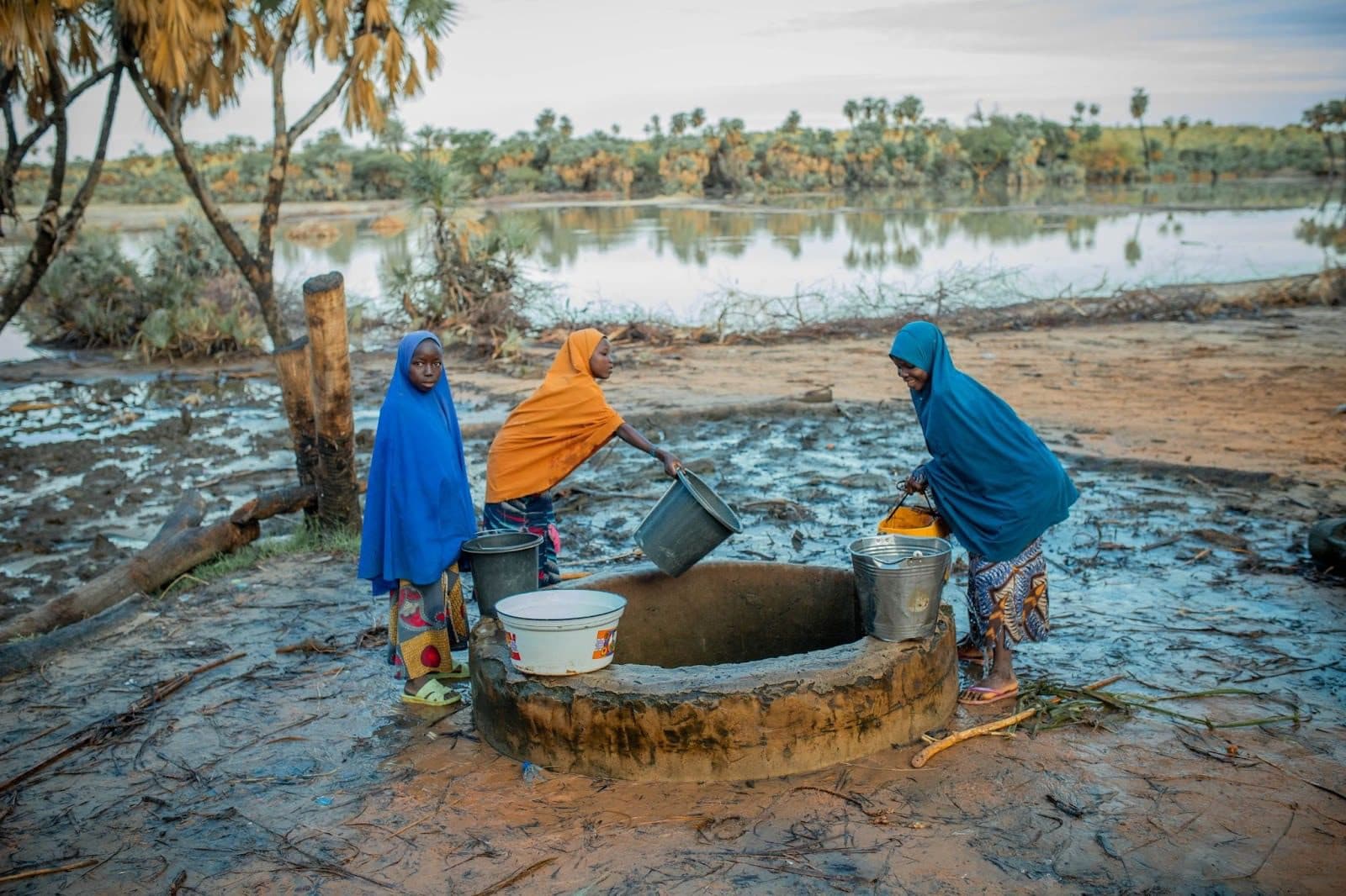 Three people gather water from a well beside a river, surrounded by trees and muddy ground.