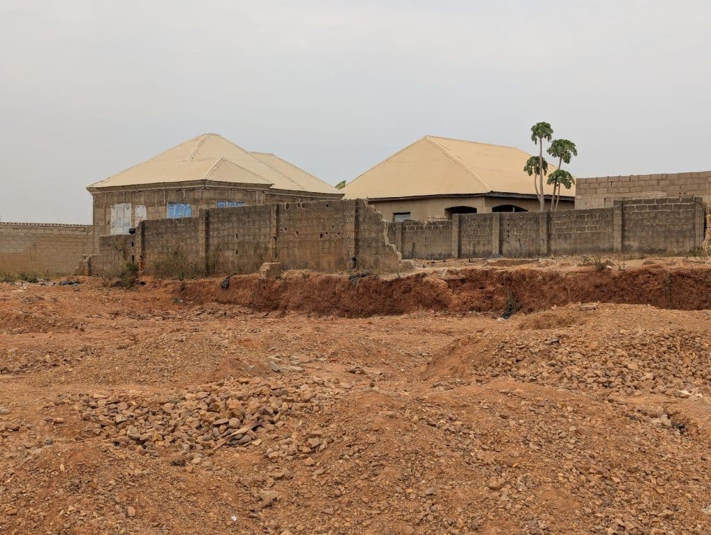 Houses with tin roofs behind a stone wall, surrounded by an empty, rocky, reddish-brown plot.