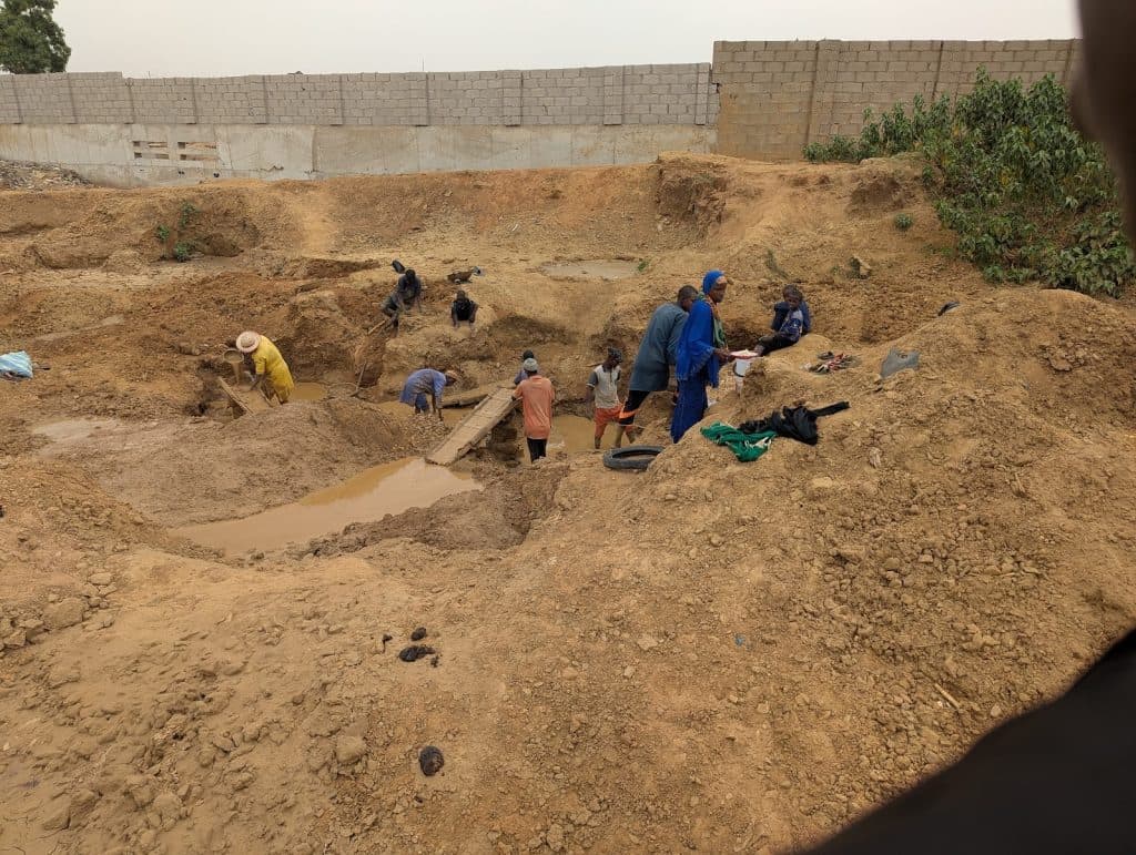 People working at a construction or excavation site, with tools and muddy water around, near a brick wall and shrubs.
