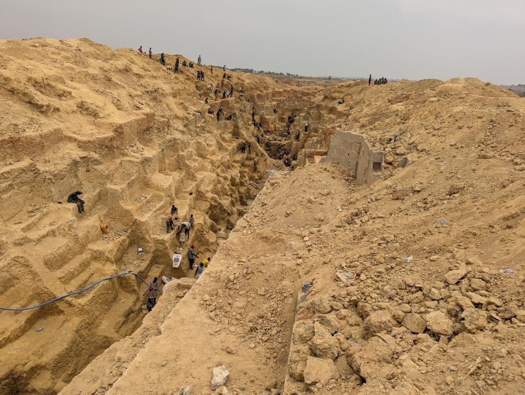 People working in a large, stepped sandy excavation site, scattered across different levels, with tools and equipment visible.