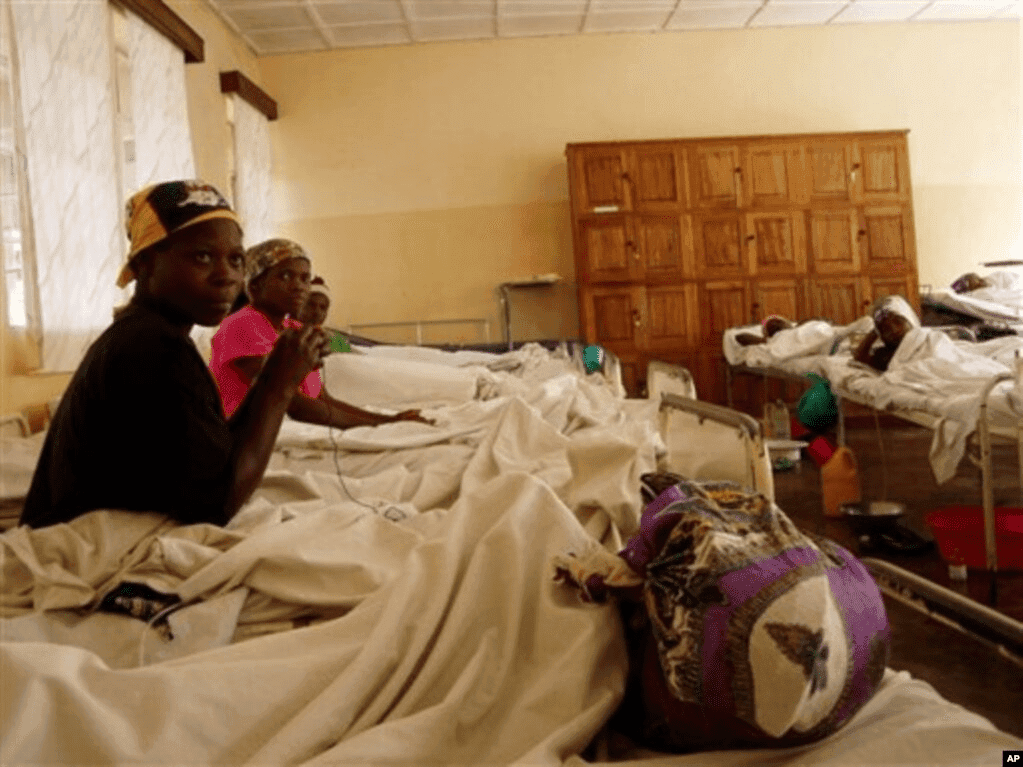 Hospital ward with several patients in beds, some sitting up, with wooden cabinets in the background.