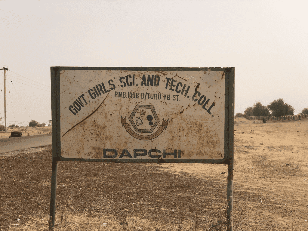Rusty sign reading "Govt Girls Sci. and Tech. Coll. Dapchi" in a dry, rural landscape.