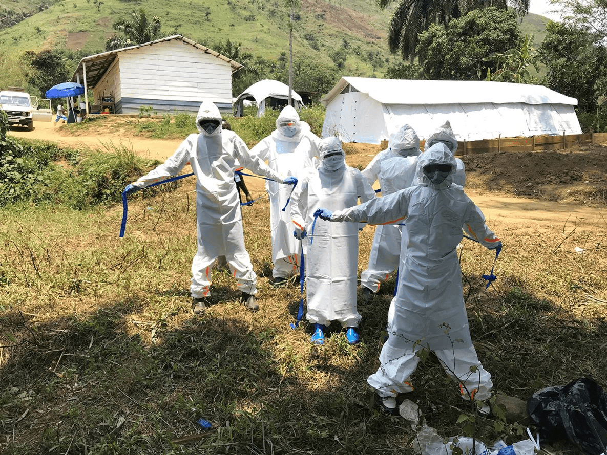 People in full protective suits, masks, and goggles standing outside near tents and a building in a rural area.