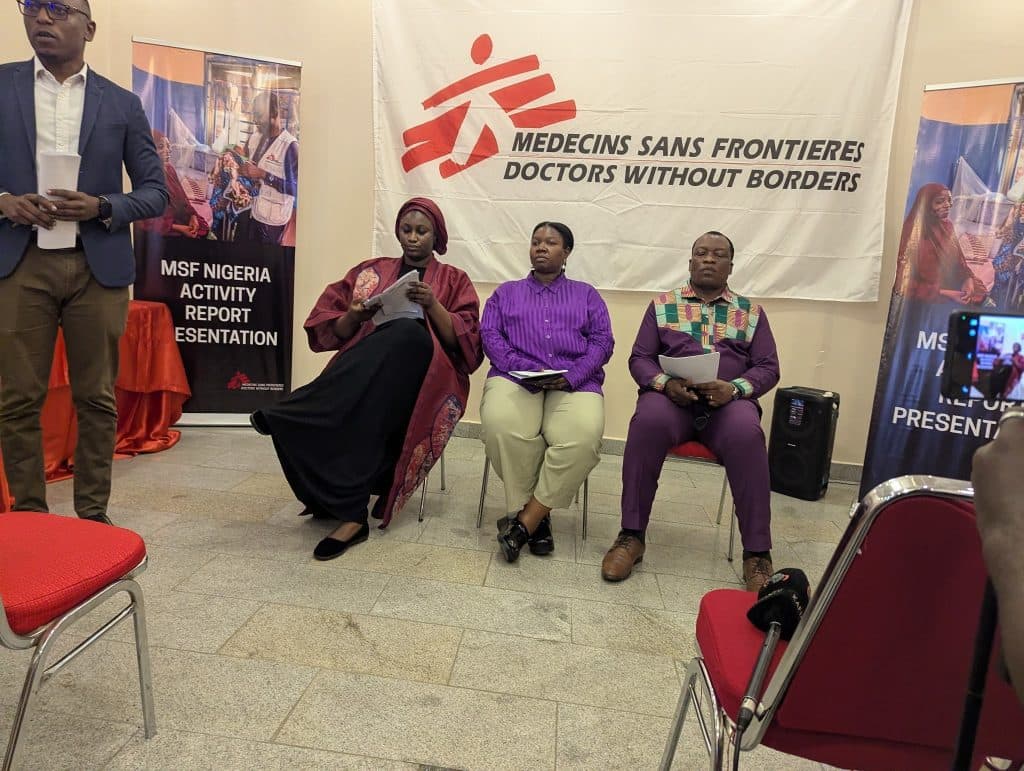 Four people in a room during the MSF Nigeria Activity Report Presentation. A banner and posters are visible in the background.