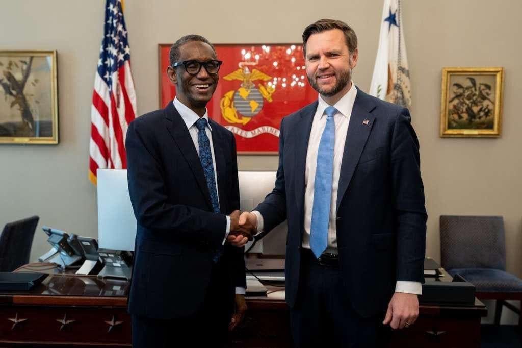 Two men in suits shake hands in an office with flags, paintings, and a desk in the background.