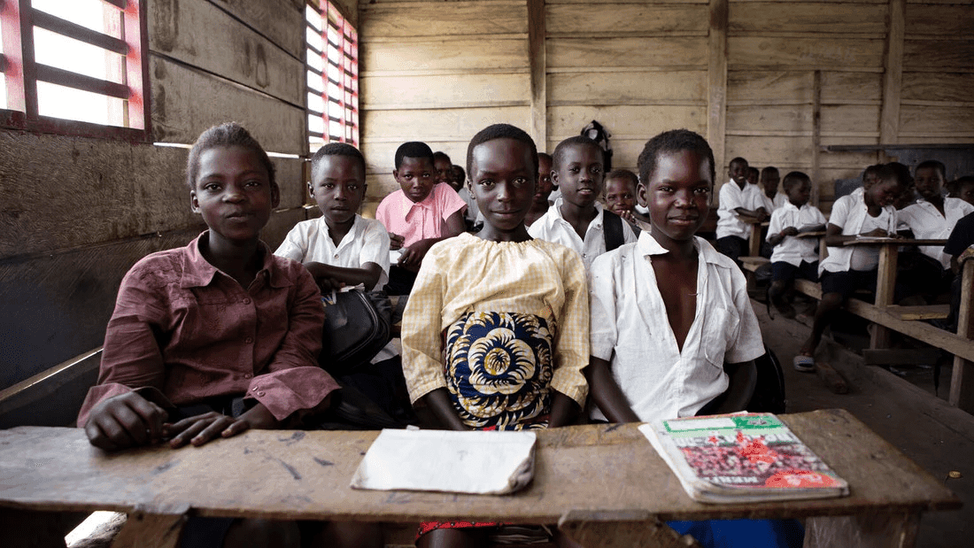 A group of children in a classroom, seated at wooden desks, looking toward the camera with notebooks in front.