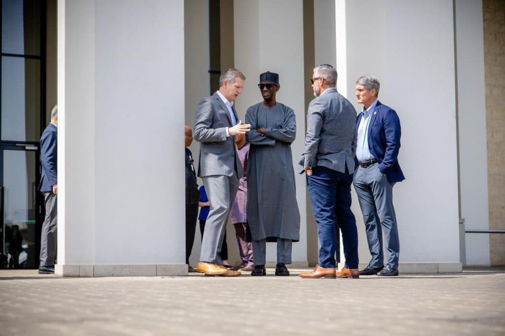 Four men in business attire stand in conversation near a white building, one in a traditional outfit.