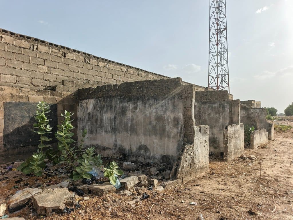 Abandoned concrete structures with green plants growing amid rubble, with a telecom tower in the background under a clear sky.