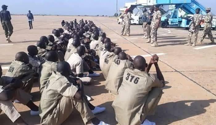 A group of men in numbered uniforms sit on the ground, facing military personnel on an airstrip.
