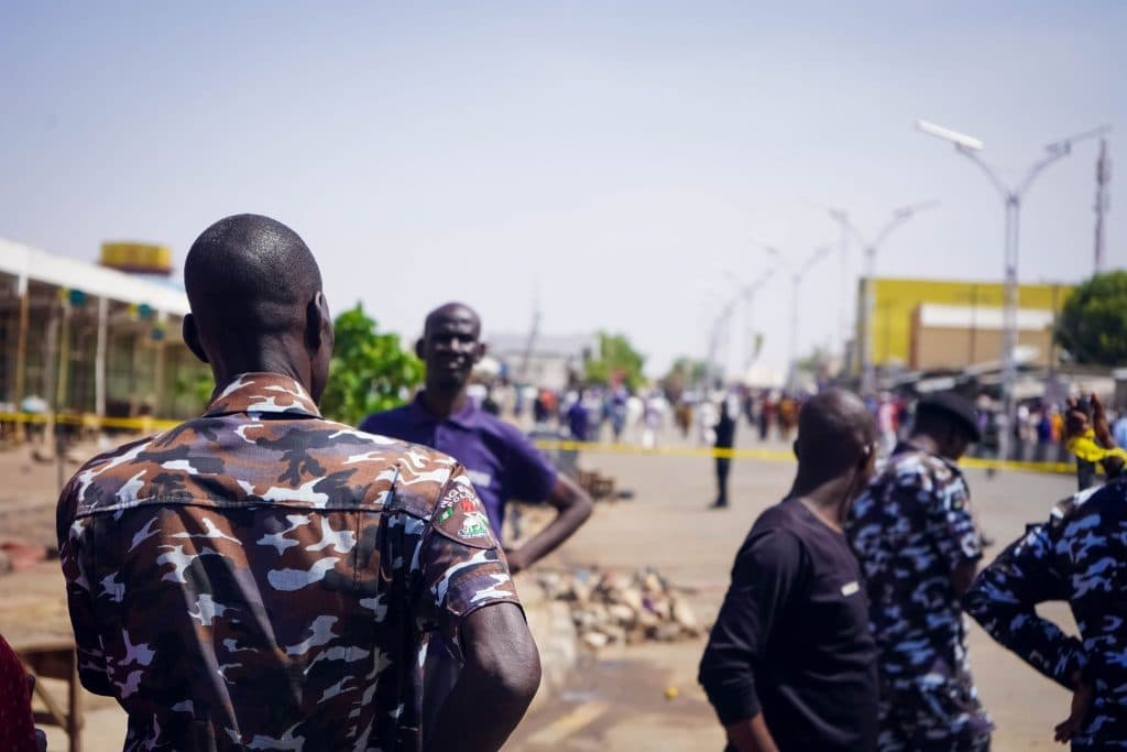 People in uniforms and civilians gather on a street, with yellow tape indicating a restricted area.