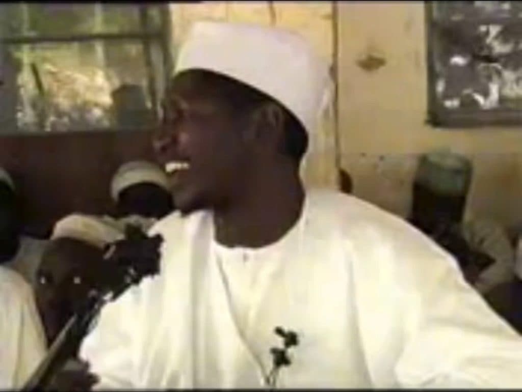 A man in white traditional attire and cap smiles, turning his head to the side. Others in similar attire are in the background.
