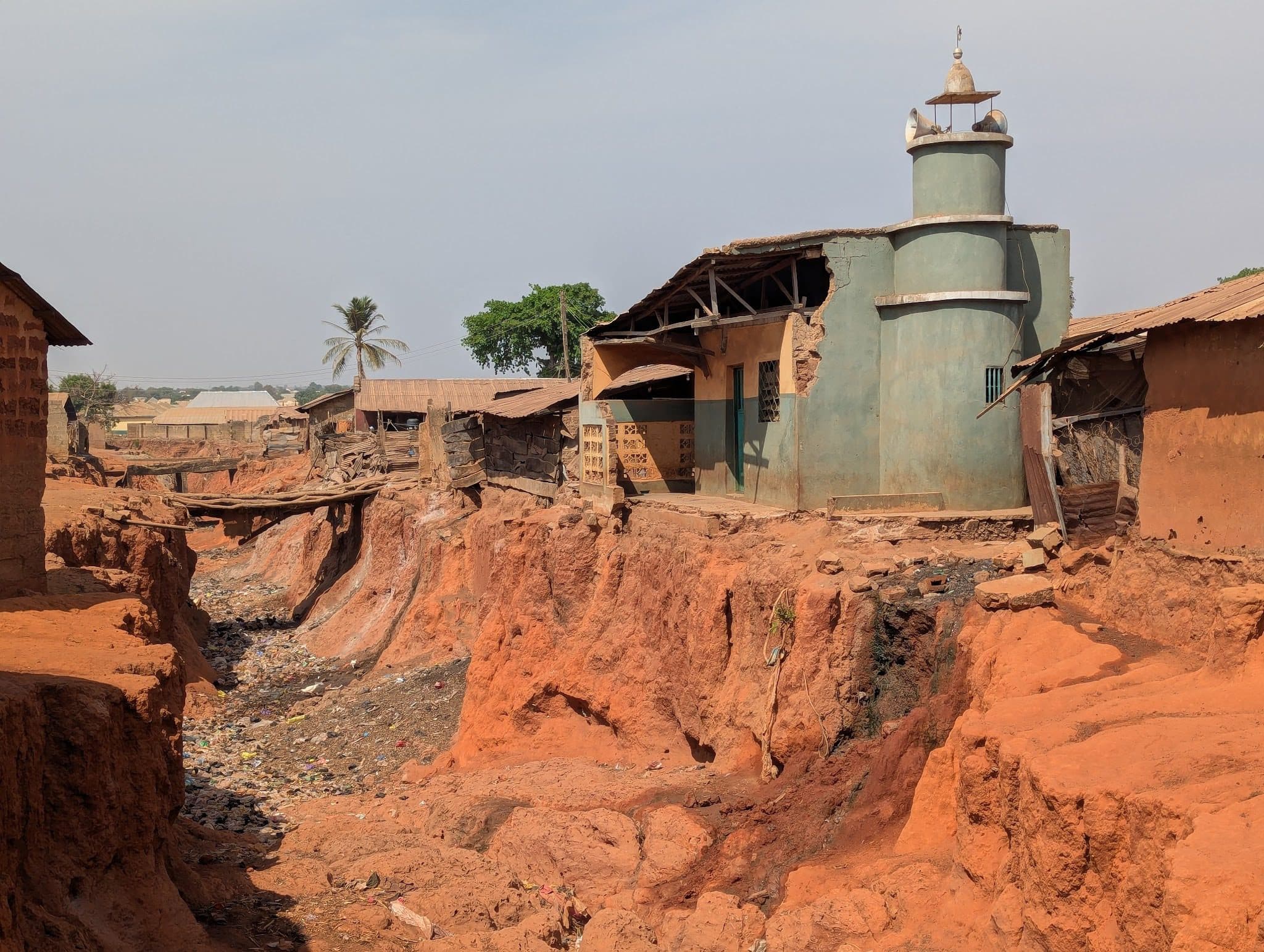 Eroded red soil surrounds a small mosque; houses and a palm tree stand nearby under a clear sky.
