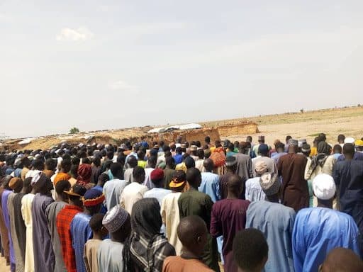 A large group of people stand in rows outdoors, appearing to observe or participate in an event under a clear sky.