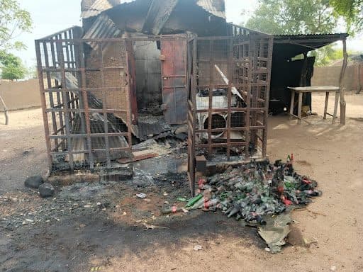Burned metal structure with charred roof, surrounded by melted glass bottles and blackened debris on the ground under sunny skies.