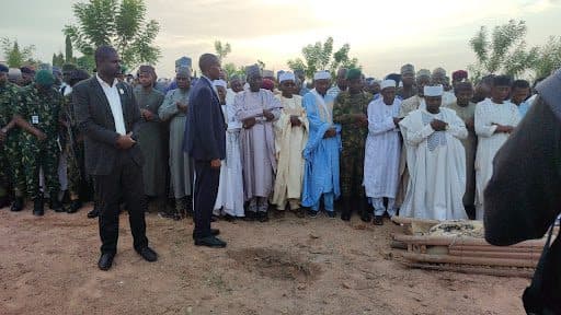 A group of people gather outdoors in traditional attire, some praying, near a covered body on a stretcher.