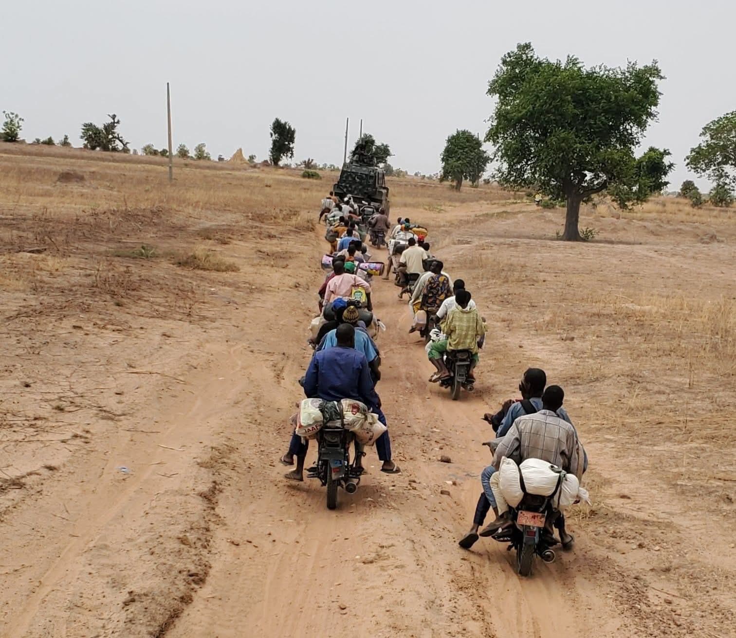 People on motorcycles follow a military vehicle on a dirt road through a dry landscape with sparse trees.