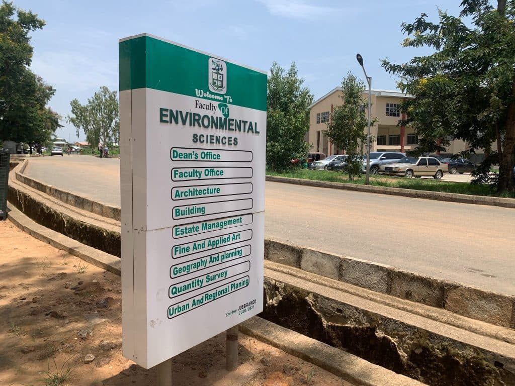 Sign for the Faculty of Environmental Sciences by a road, listing offices like Architecture and Geography. Trees and a building in the background.