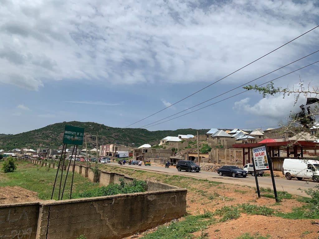 Street view with a mountain backdrop, signs for a university hostel and gas plant, parked cars, and buildings under a cloudy sky.