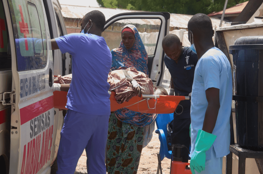 Medical personnel load a patient onto an ambulance in a rural area, while a woman stands nearby.