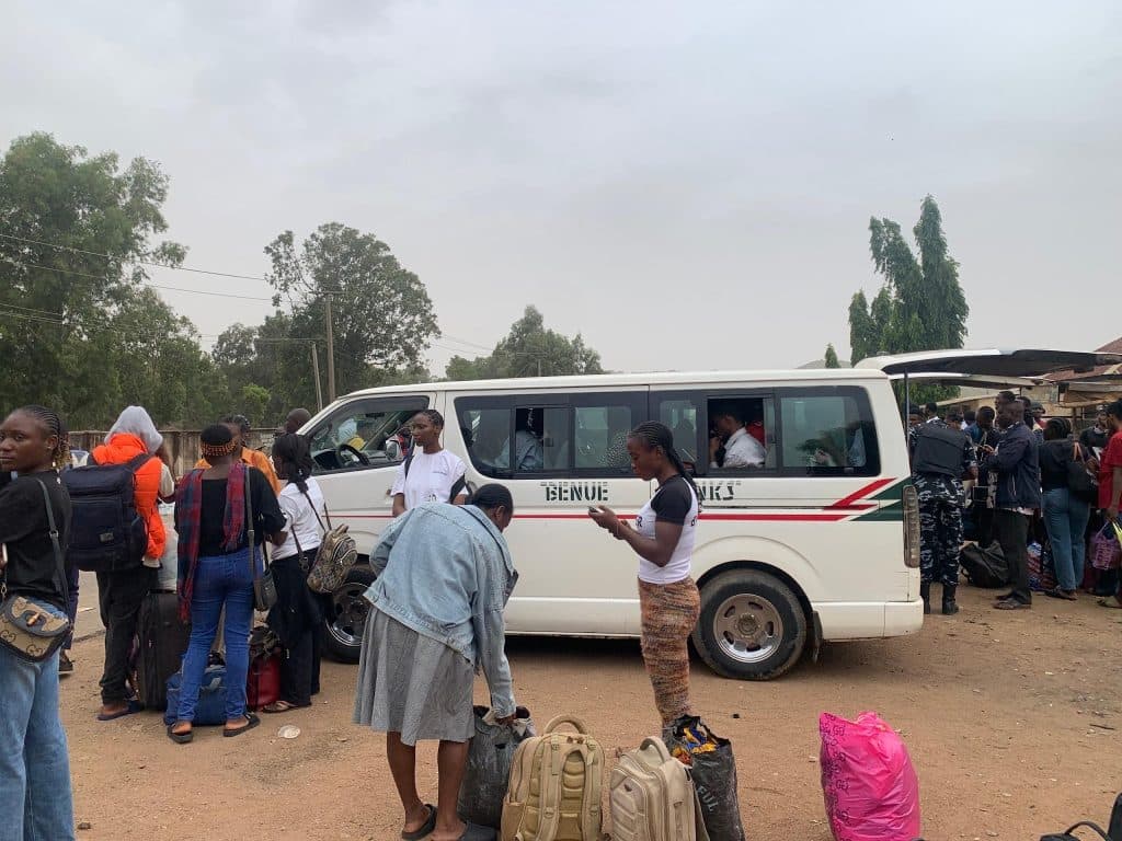 People standing with luggage near a white minibus labeled "Benue Links" on a dirt road, surrounded by trees and overcast sky.