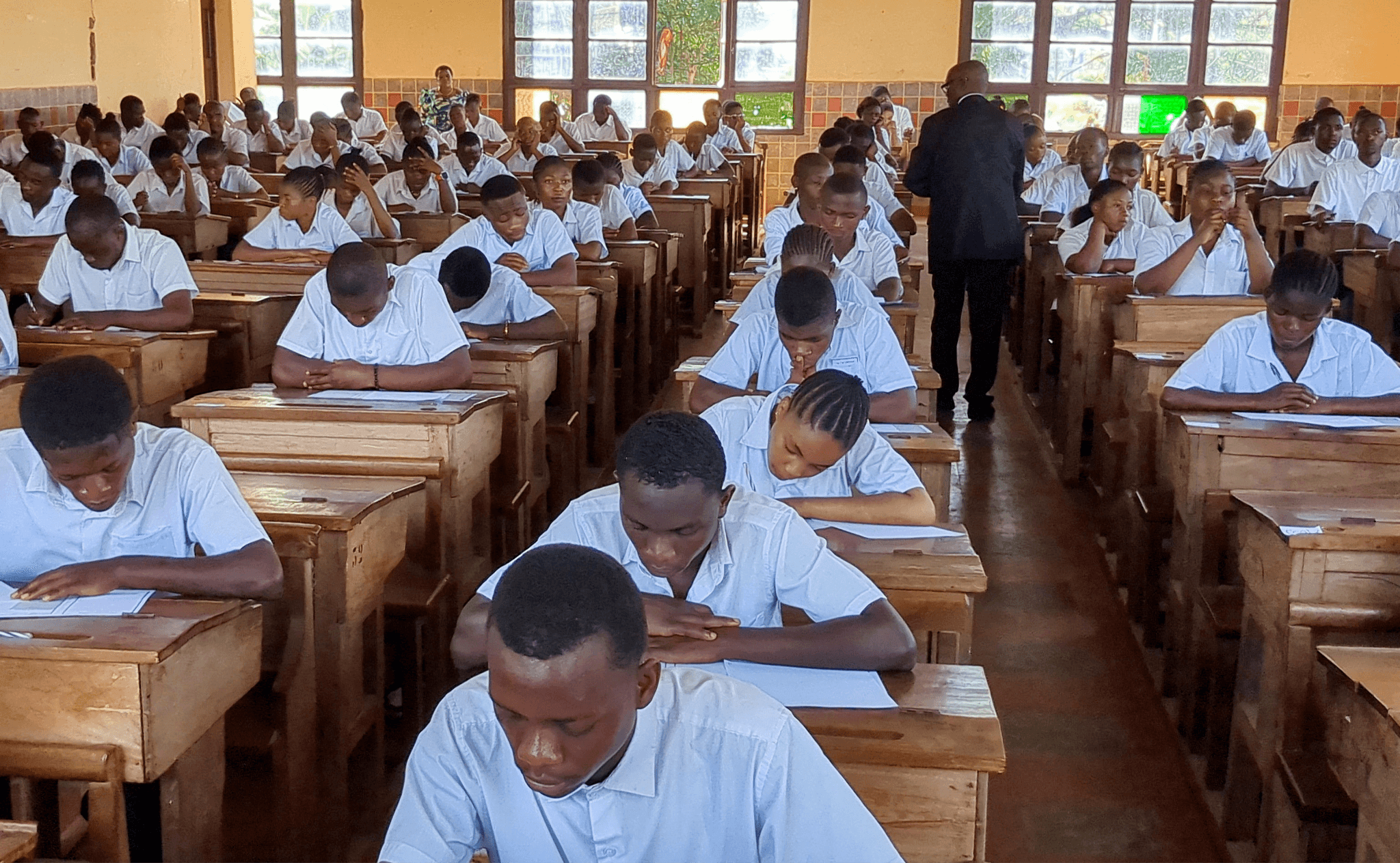 Students in uniforms sitting in rows at wooden desks, focused on taking an exam in a classroom setting.