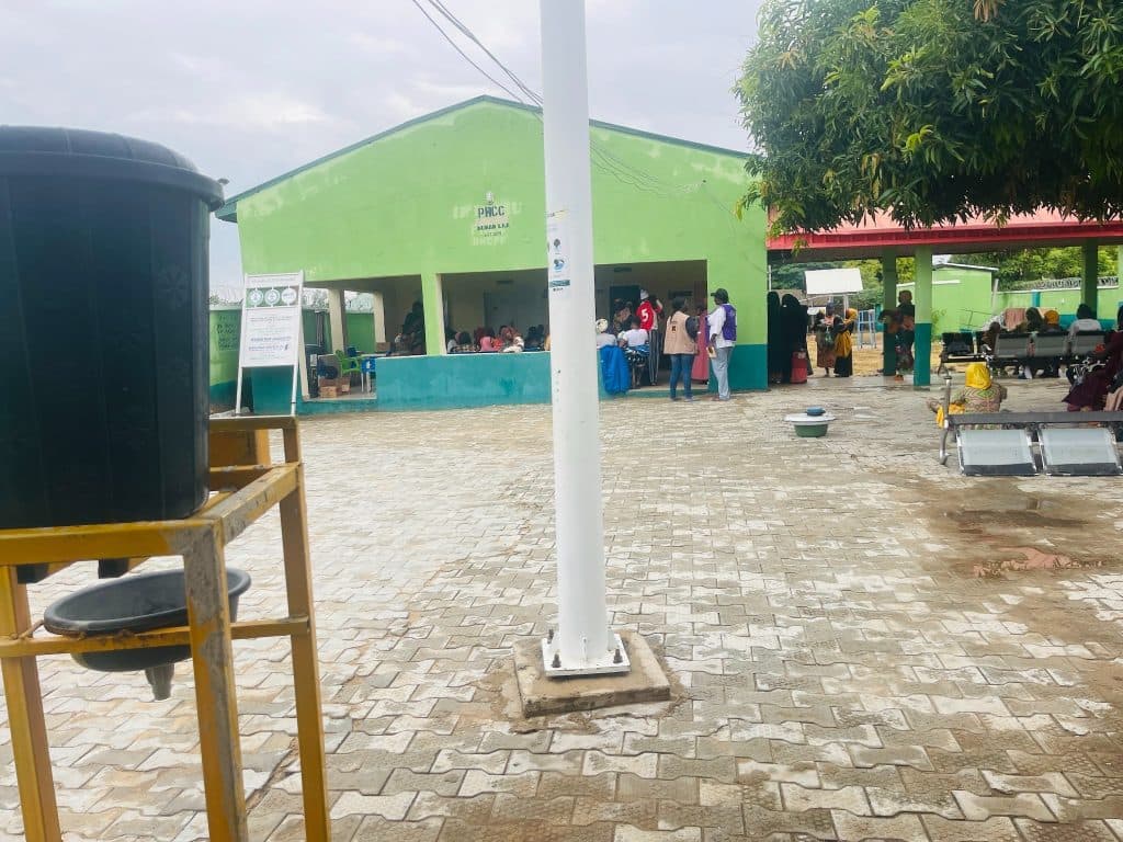 Outdoor scene with people gathered at a green building, likely a health or community center. A handwashing station is visible in the foreground.