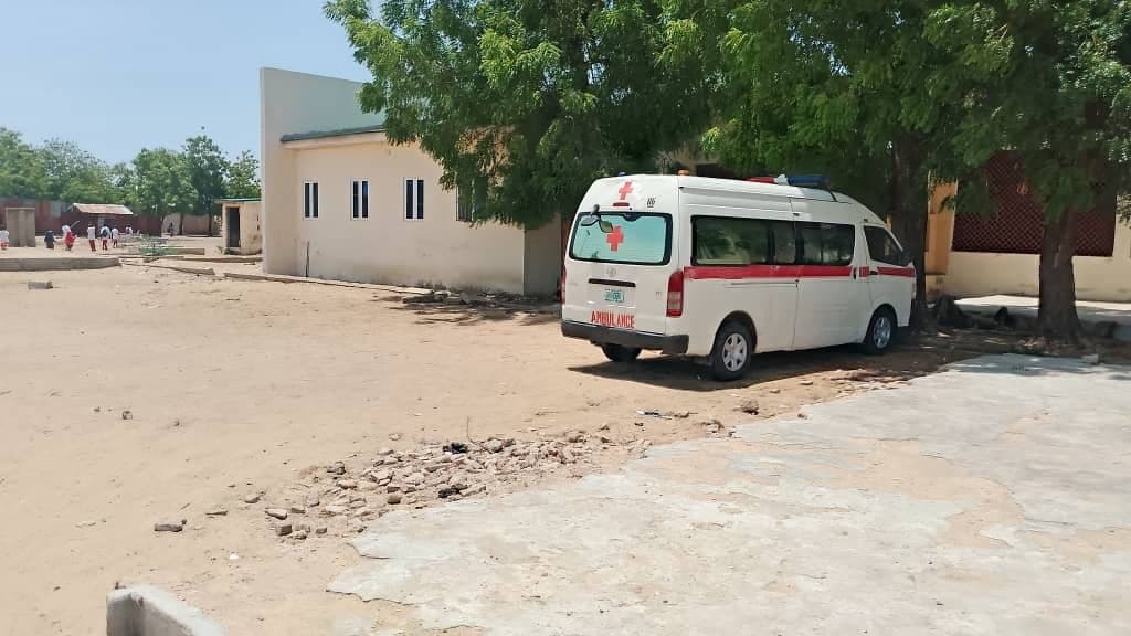 An ambulance is parked under a tree near a light-colored building in a sandy area on a sunny day.