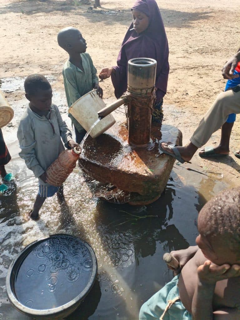 Children collecting water at a hand pump in a rural area, with containers and a basin.