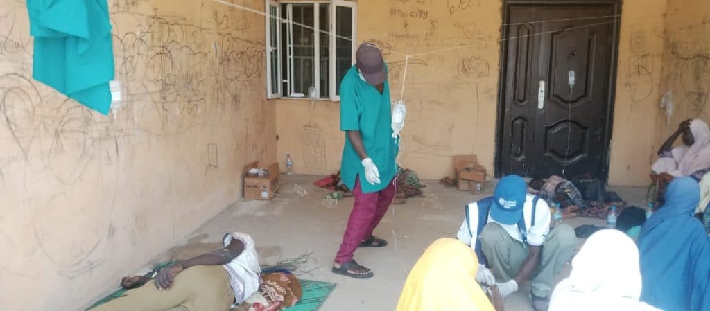 People receiving medical treatment in an outdoor area, with healthcare workers attending to them. Walls show graffiti and a door is visible.