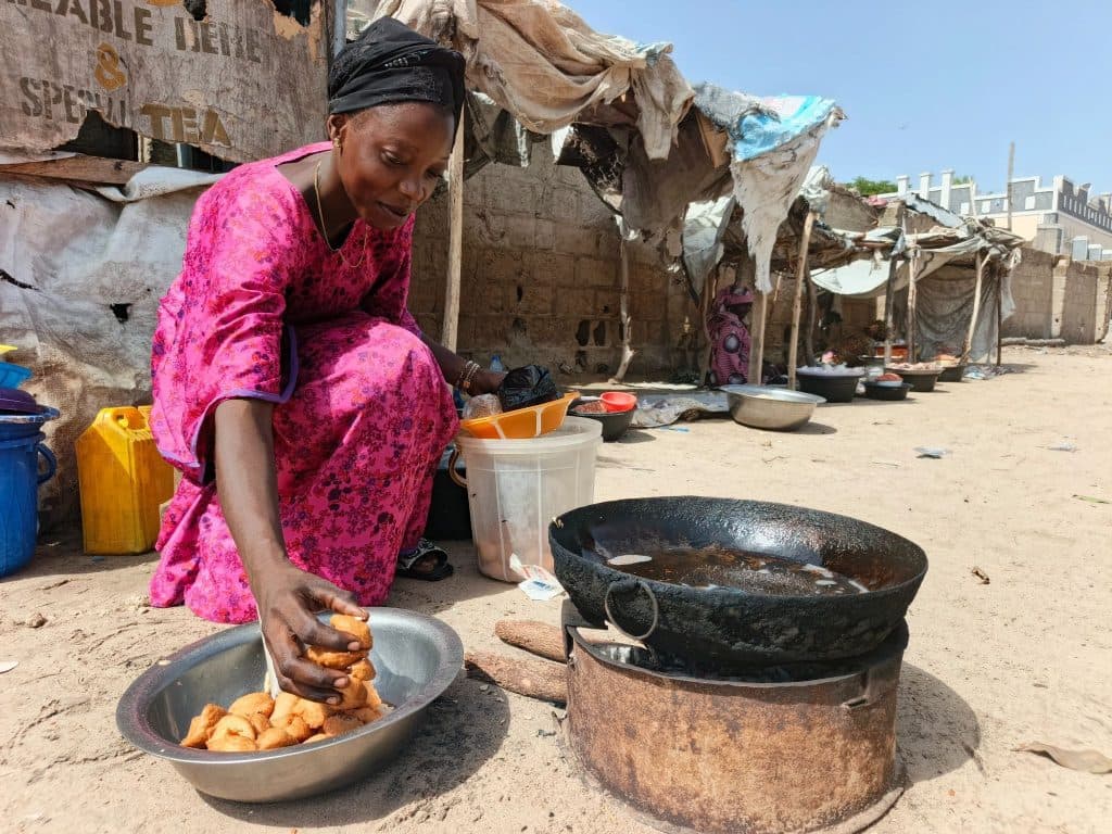Woman in pink dress cooking by an outdoor stove, placing food into a bowl, with tents and cooking supplies in the background.