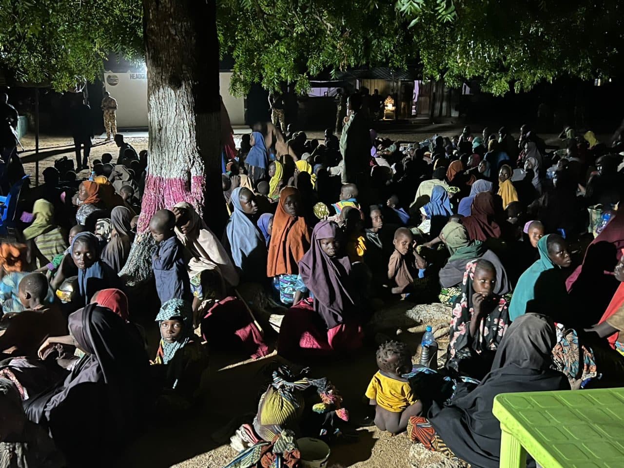 A large group of people, including children, sitting under a tree at night. Many are wearing colorful clothing and head coverings.