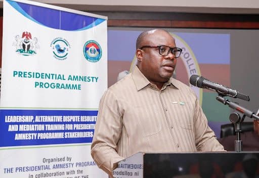 Man speaking at a podium during the Presidential Amnesty Programme event. A banner with program details is visible behind him.
