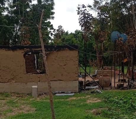 A partially burned building with bare walls and charred remains in a grassy area, surrounded by trees.