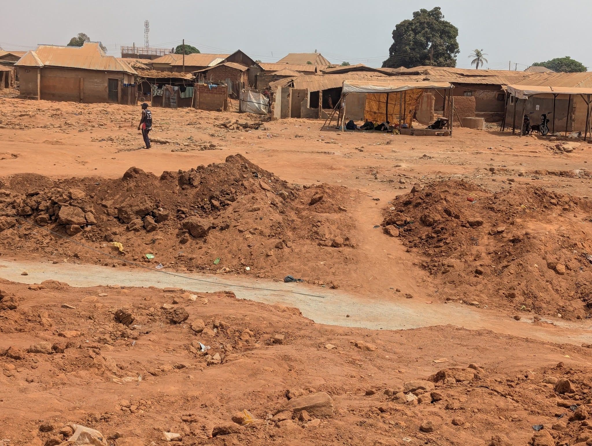 A lone person walks across a dry, rugged, reddish-brown landscape with a background of rustic buildings under a clear sky.