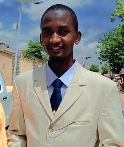Smiling person in a light suit and tie outdoors, with trees and a bright sky in the background.