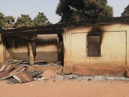 Burned building with charred walls, damaged roof, and debris on the ground surrounded by trees.