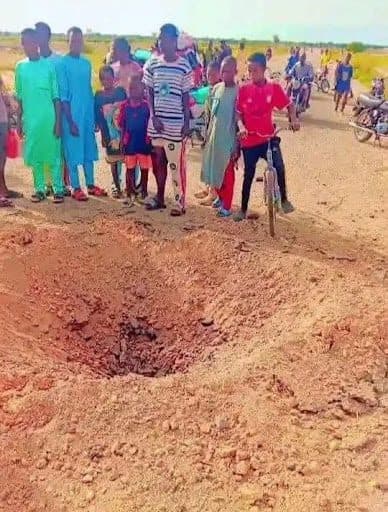 Group of people and children with bikes gathered around a large crater on a dirt road.